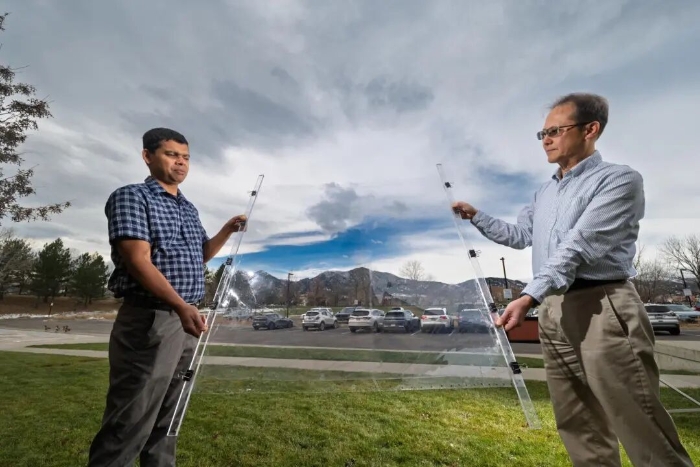 Two men hold a large sheet of clear material in between them with mountains visible in the background